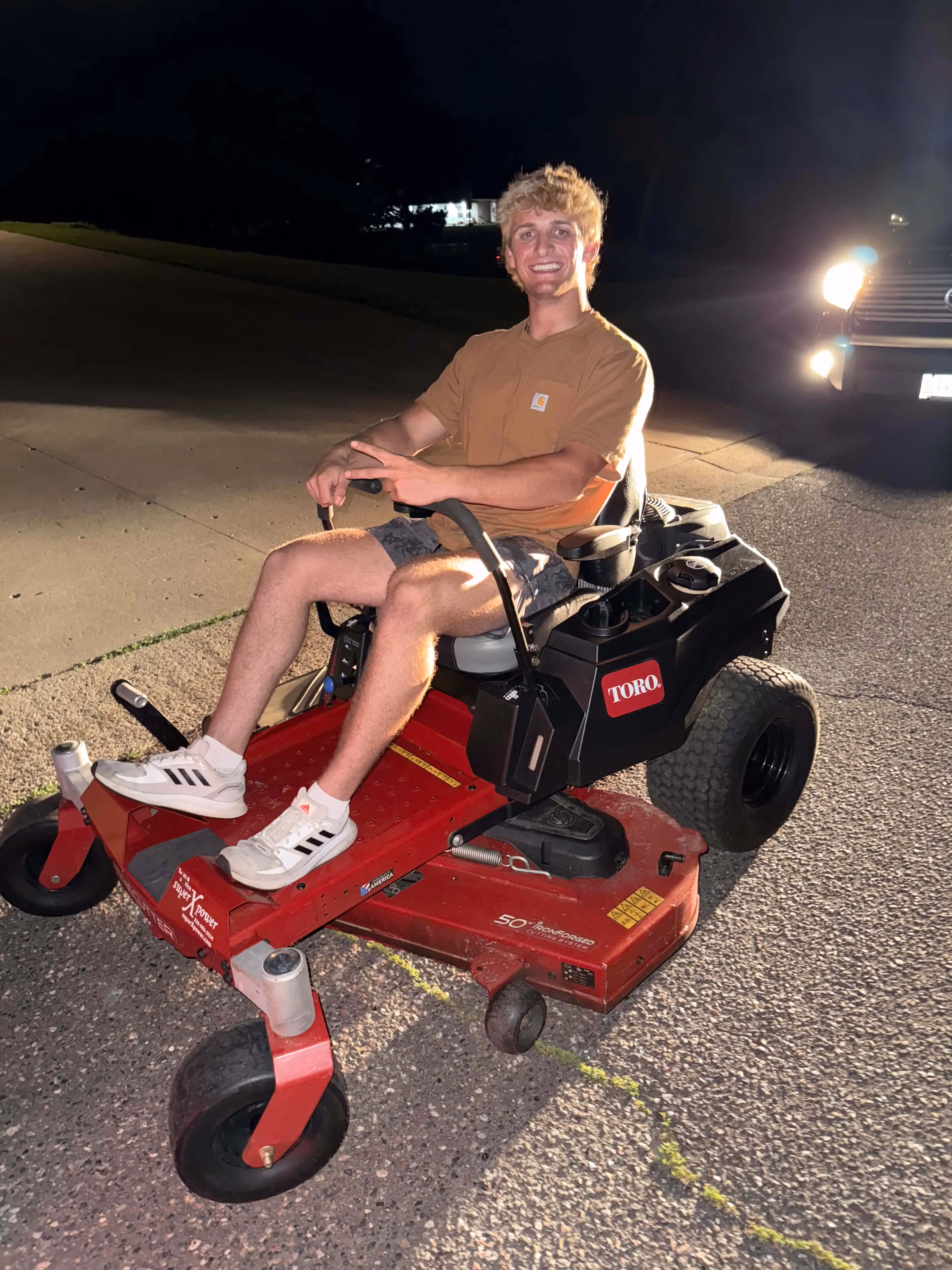 Smiling young man in brown shirt sitting on a red Toro zero-turn riding lawn mower at night.