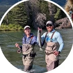 A man and woman standing in a river holding fishing rods and a large fish smiling at the camera.