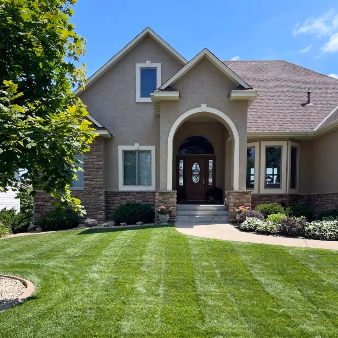Front yard with striped green lawn, shrubs, and a beige house entrance with arched doorway and wooden door.