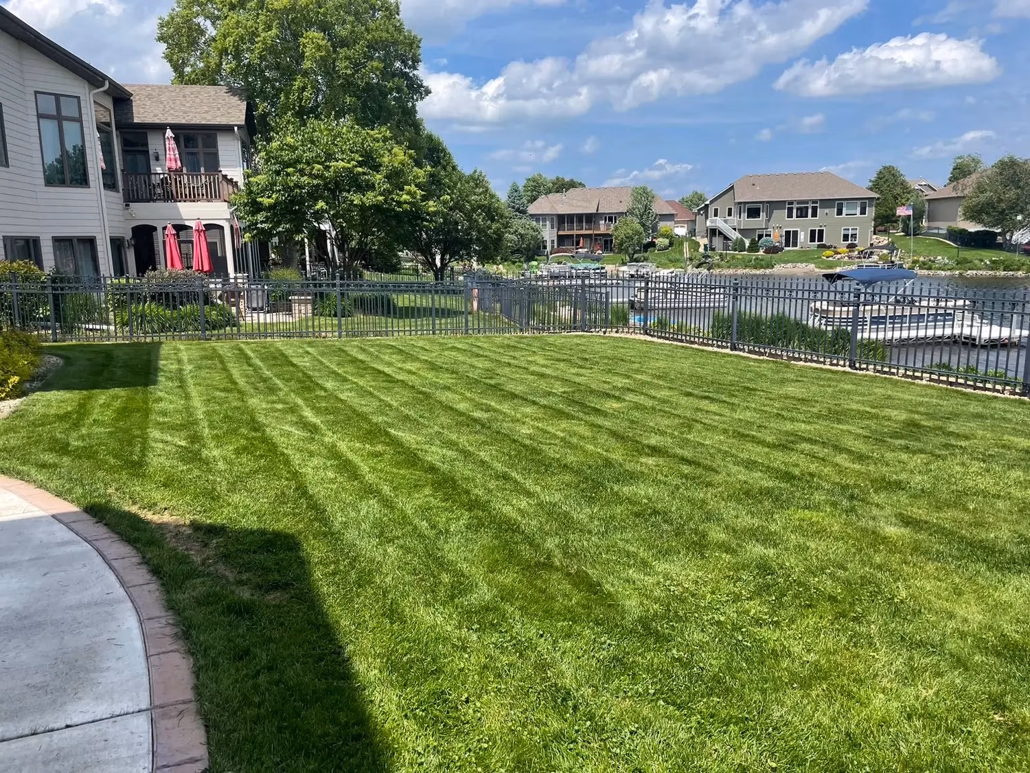 Well-maintained green lawn with striped mowing pattern by a black metal fence near a lake and houses.